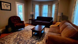 Un salon avec un canapé marron foncé, un fauteuil rouge, une table basse en bois et un tapis à motifs. Un plan encadré est accroché au mur et une lampe se trouve sur une table d’appoint.