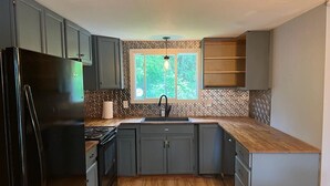 A compact kitchen with grey cabinets, a black refrigerator, a wooden countertop, and a window with a view of greenery. - Welcome to Blueberry Cottage, a peaceful Oasis (Mississippi Mills)