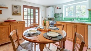 A dining area with a wooden table set for two, a kitchen with green tiles, and a large window providing natural light. - Les Tourelles by Interhome (Dinard)