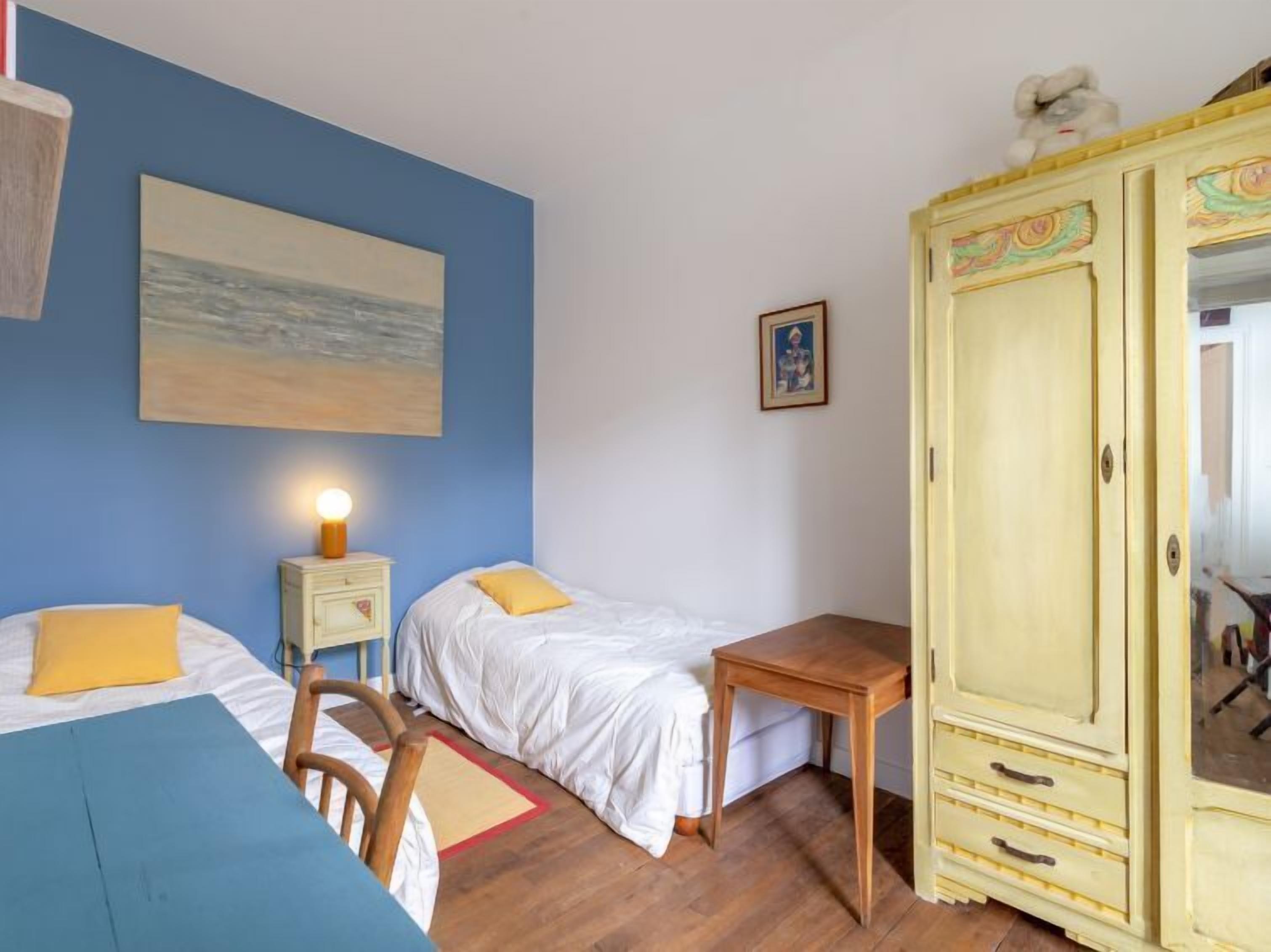 Une chambre avec deux lits, une table en bois, une table de chevet avec une lampe, une armoire et un tableau au mur.