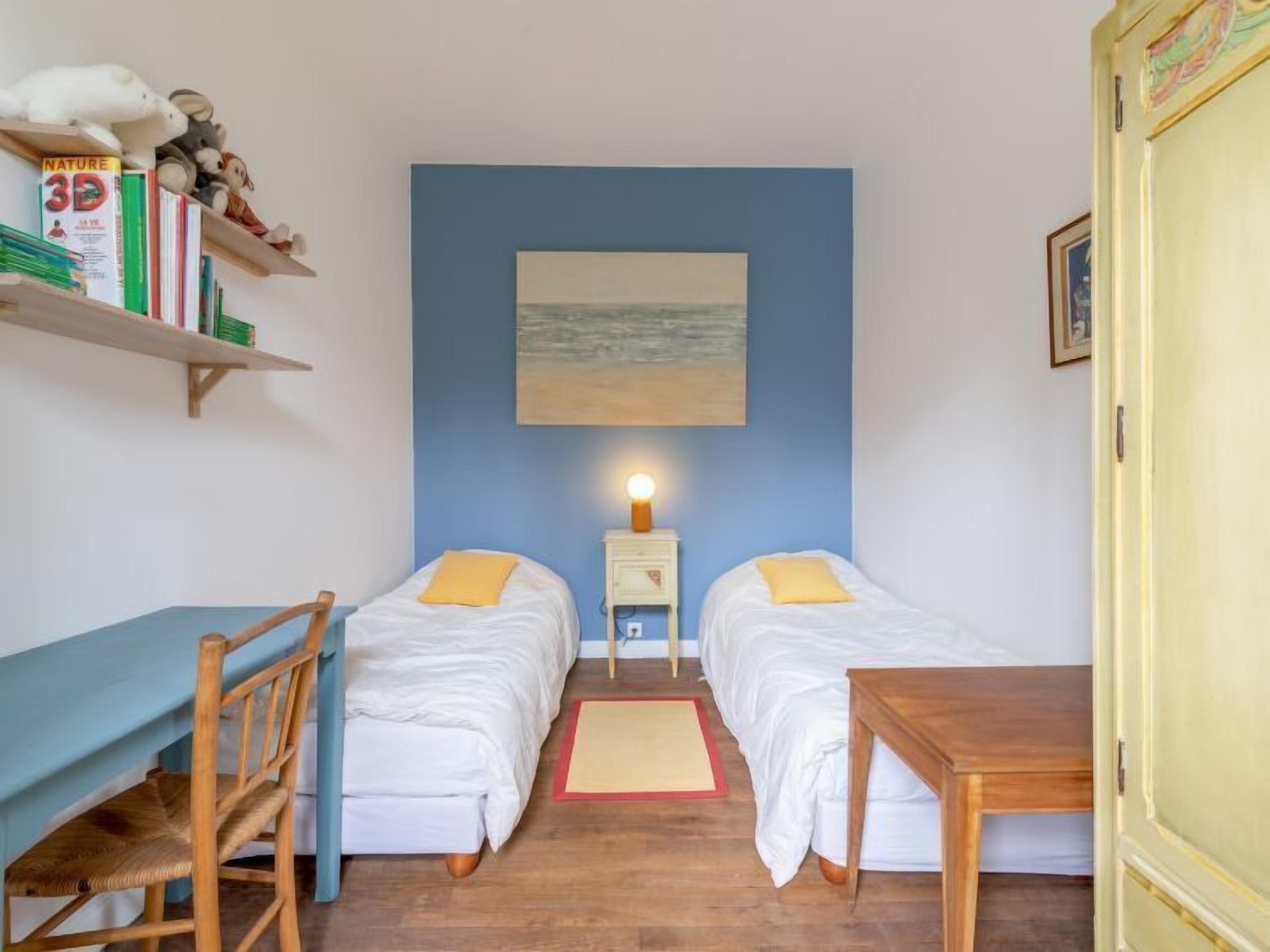 A room with two beds, a desk, a chair, a shelf with books, and a painting on the wall.
