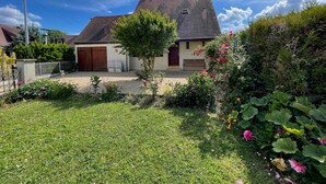 Une maison à deux étages, dotée d’un toit en pente, d’un porche à l’avant et d’un jardin bien entretenu où poussent diverses plantes et fleurs.