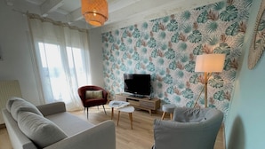 Un salon comprenant un canapé, un fauteuil, un téléviseur et un mur recouvert d’un papier peint à motifs de feuilles tropicales.