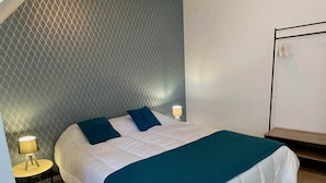 Un lit bien fait, avec une literie blanche et des touches de couleur turquoise, une table d’appoint ronde avec une lampe, et un mur orné d’un motif géométrique.