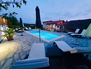 Una piscina de piedra con sillas de descanso y sombrillas, rodeada de edificios con techos de tejas rojas.