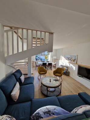 Uma sala de estar moderna com um sofá azul, duas poltronas amarelas, uma mesa de centro redonda e uma escada que dá para outro andar.