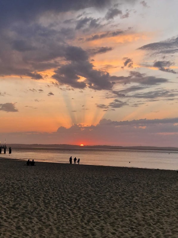Uma praia ao pôr do sol, com céu claro, mar calmo e um píer ao longe.