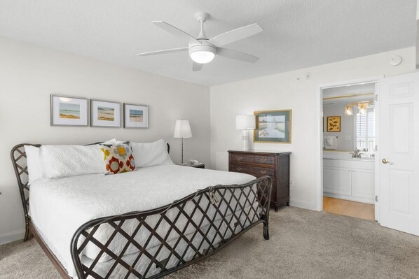 Un dormitorio con cama, ventilador de techo, cómoda, mesita de noche y puerta que da al baño.