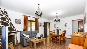 A living room with a gray sofa, wooden coffee table, and a dining area with a wooden table and chairs. There is a fireplace, a chandelier, and a stone wall. - 014 Villa Colonia (Denia)