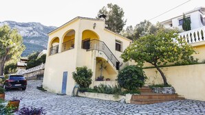 A yellow building with a staircase leading to a balcony, surrounded by greenery and a cobblestone street. - 014 Villa Colonia (Denia)