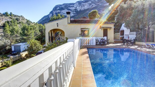 A poolside area with a clear view of a mountain, a building with a tiled roof, and a white railing. - 014 Villa Colonia (Denia)