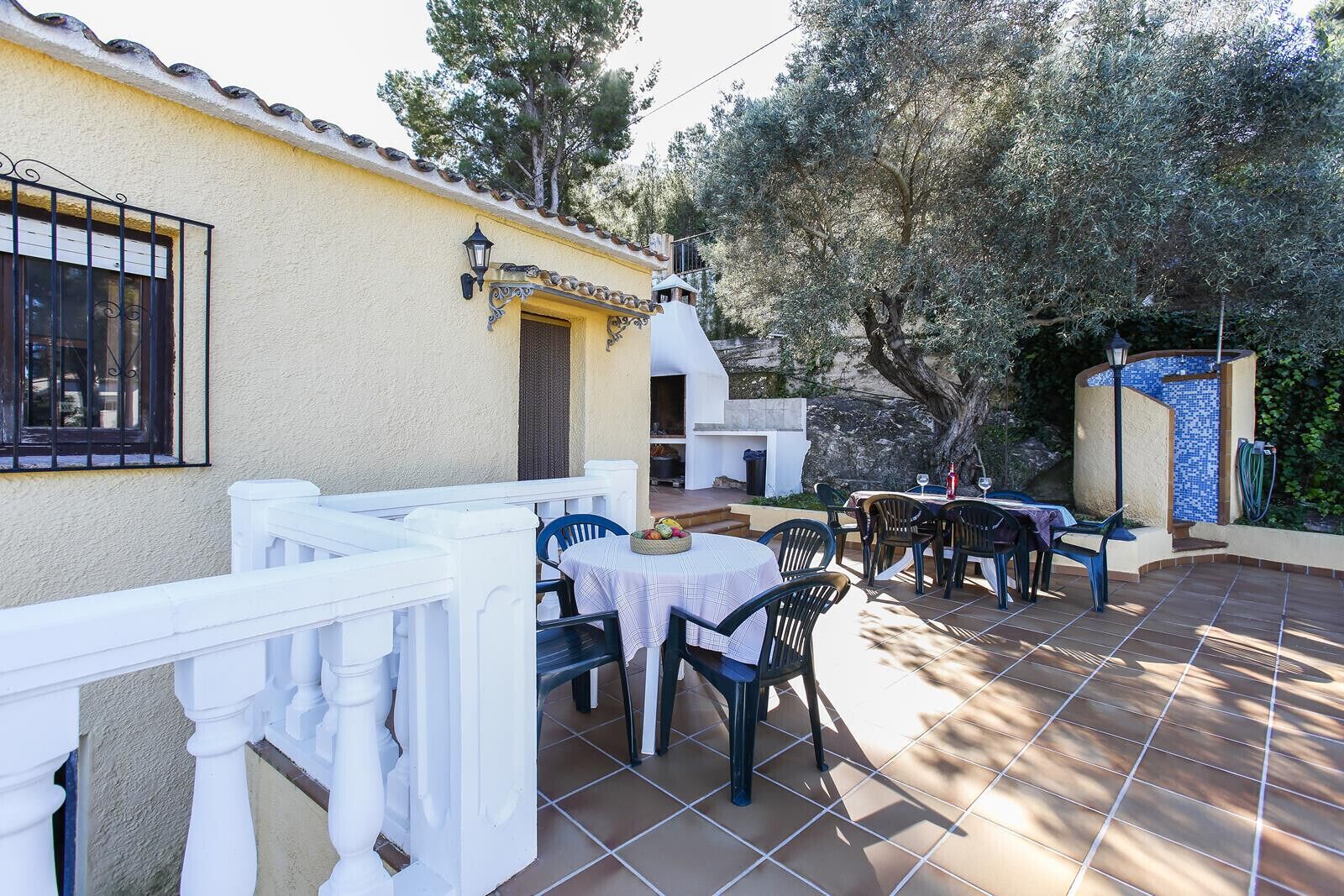 Une terrasse extérieure avec des tables et des chaises blanches, un sol carrelé et un mur en pierre orné d’une mosaïque.