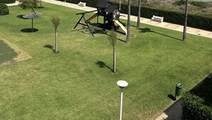 Un césped cuidado con un parque infantil, palmeras y vista al mar.