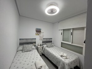 Slaapkamer met twee bedden, een nachtkastje, een wandplank met decoratieve objecten en een raam met jaloezieën.