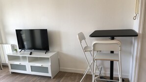Um quarto pequeno com um suporte branco para TV, uma cadeira dobrável e uma mesinha.