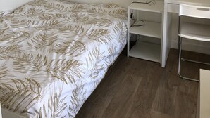 Quarto pequeno e bem arrumado, com uma cama, uma escrivaninha e uma luminária.