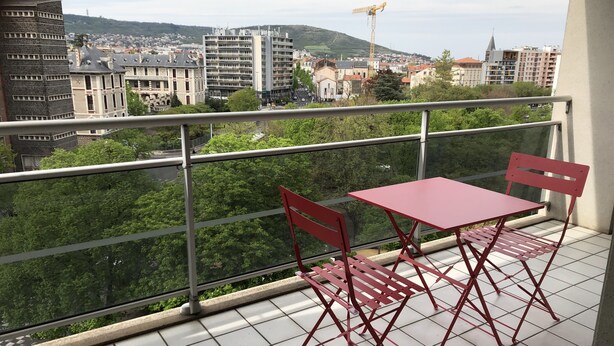Varanda com mesa e cadeiras vermelhas, exibindo vista para um horizonte urbano com edifícios e árvores.