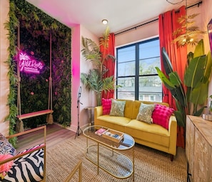 A modern living room with a yellow sofa, a glass coffee table, a neon sign, and a large window.