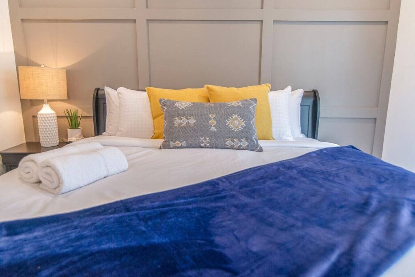 A neatly made bed with a blue blanket, white and yellow pillows, and a rolled white towel on the side.