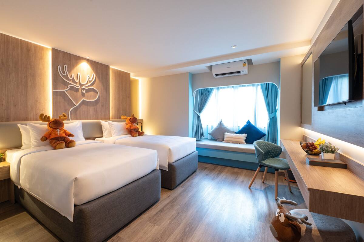 A modern hotel room with two beds, a seating area with a view, and a small desk with a plant and fruit bowl.