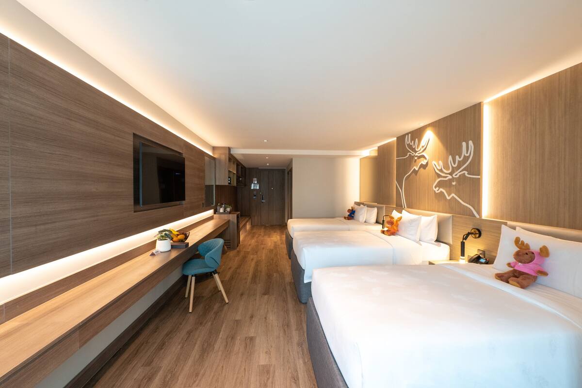 A modern hotel room with a long wooden desk, a flat-screen TV, two beds, a desk chair, and a wall-mounted artwork of reindeer.