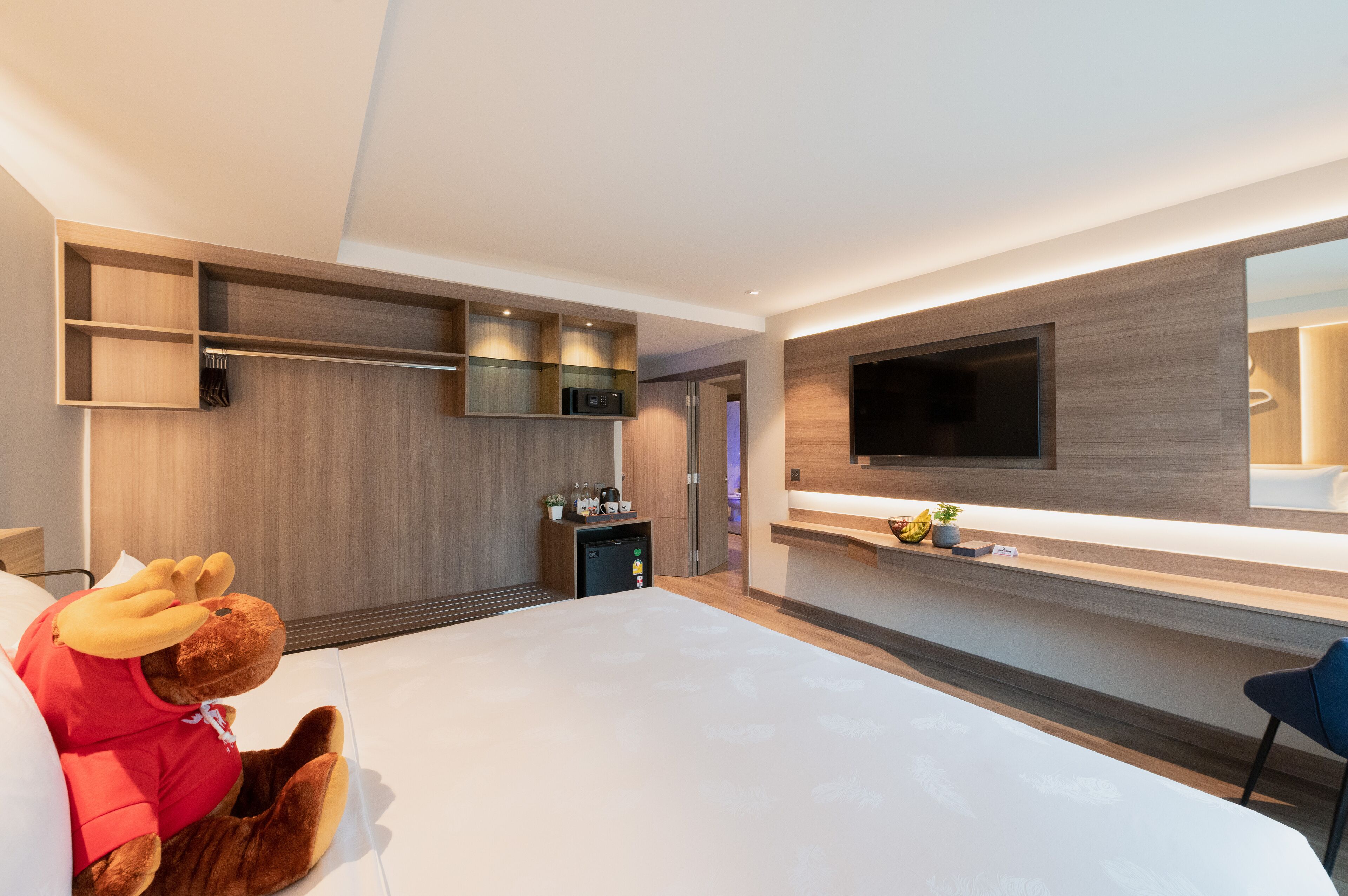 Habitación de hotel moderna con una cama grande, un televisor de pantalla plana en la pared, una pequeña kitchenette y un peluche en la cama.