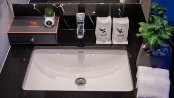 A hotel bathroom with a white sink, a glass shower, and a black countertop. There are two potted plants, a white towel, and two bags labeled "Moose Hotel."