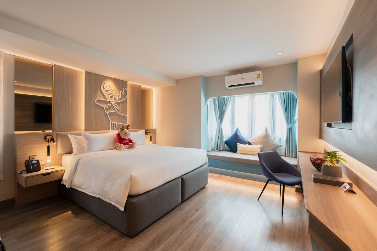 A modern hotel room with a large bed, bedside tables, a desk, a chair, and a window with curtains.