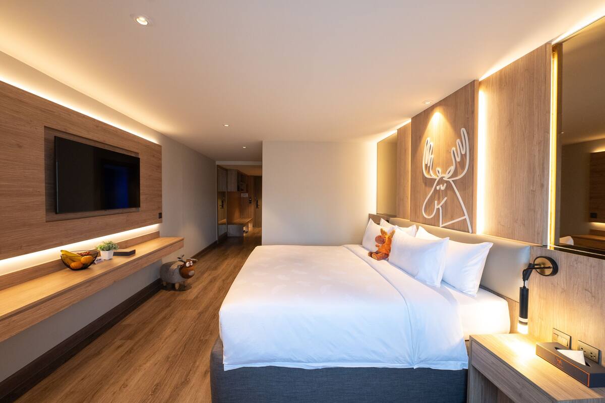 A modern hotel room with a large bed, a flat-screen TV mounted on the wall, a wooden nightstand, and a bedside table with a lamp.