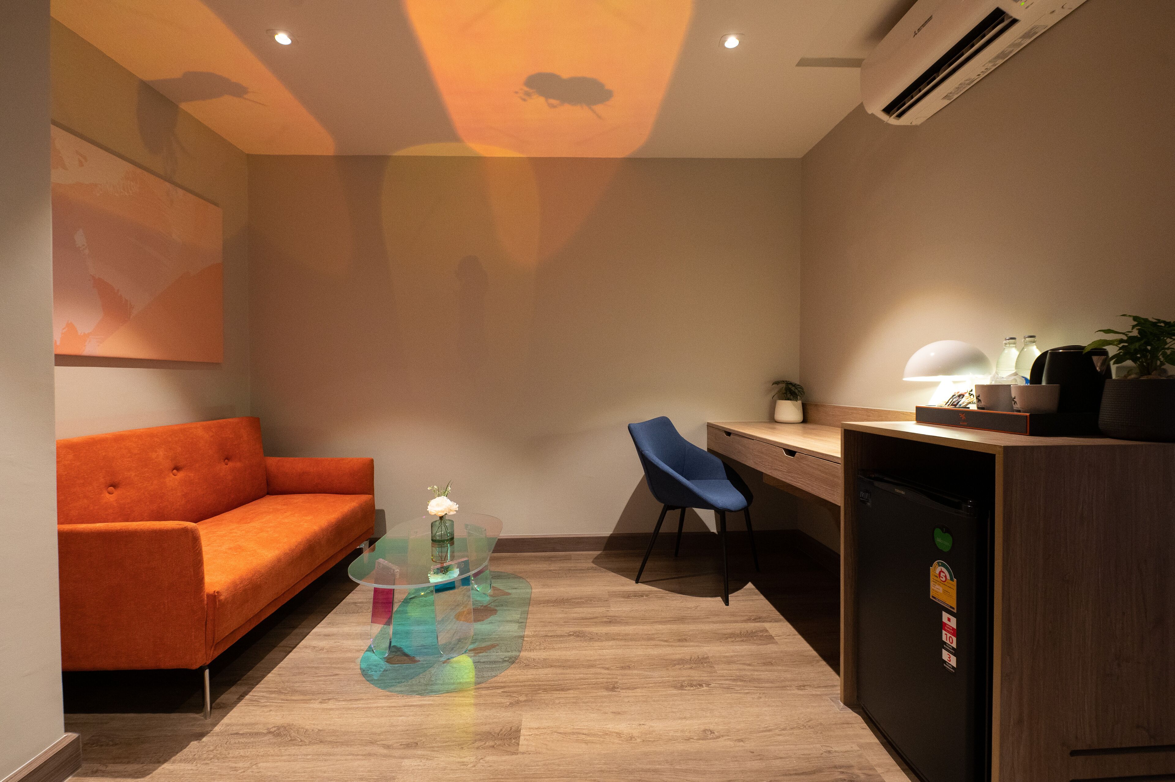 A modern room with a sofa, a glass table with a vase, a desk with a chair, a refrigerator, and a wall-mounted air conditioner.