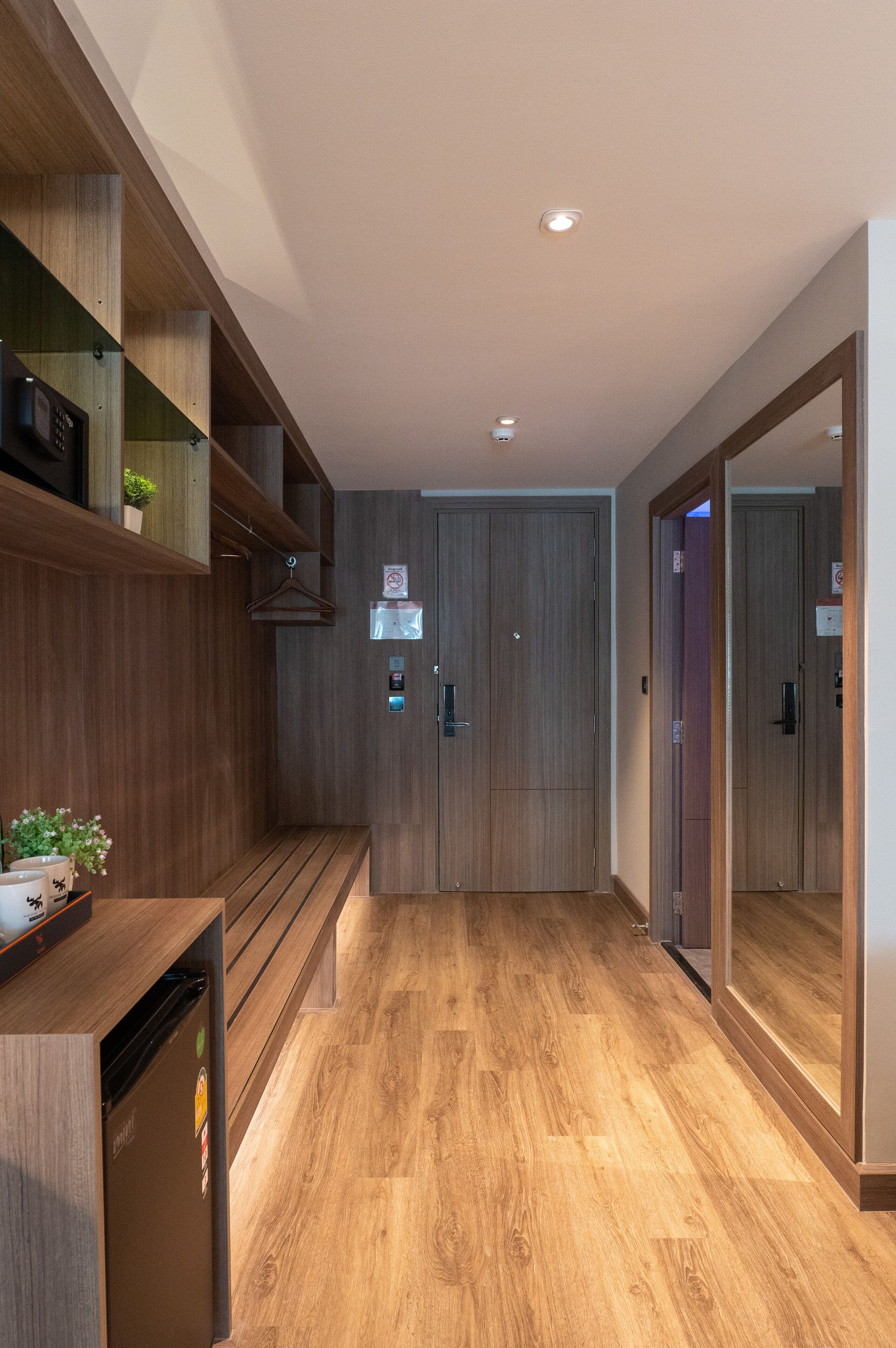 A hallway with wooden flooring, a bench, a refrigerator, and a mirror.