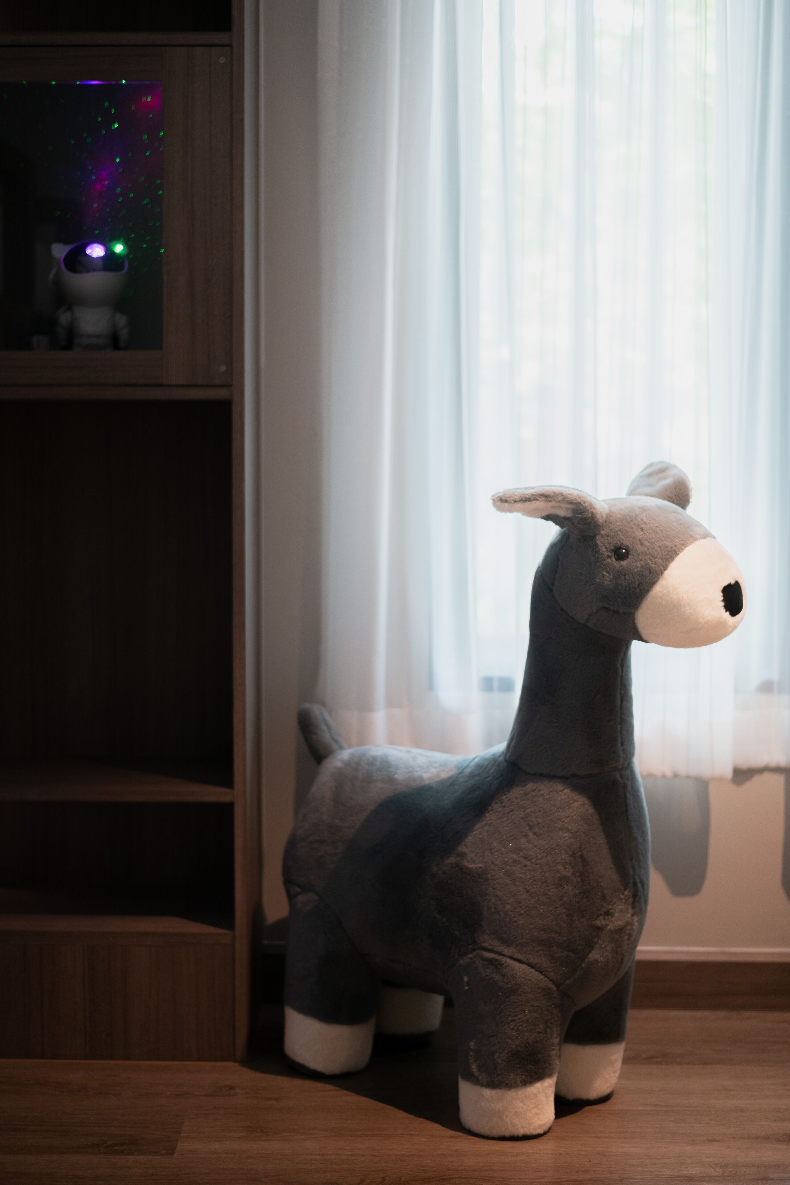 A plush toy donkey in a room with a wooden floor and a window with sheer curtains.