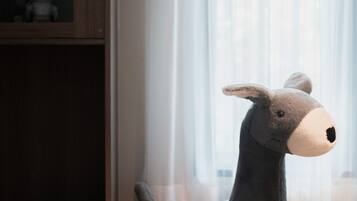 A plush toy donkey in a room with a wooden floor and a window with sheer curtains.