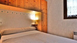 Una habitación con paneles de madera, una cama, una mesita de noche, una lámpara de pared y una ventana con cortinas de encaje.