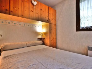 A wooden-paneled room with a bed, a nightstand, a wall-mounted lamp, and a window with lace curtains.