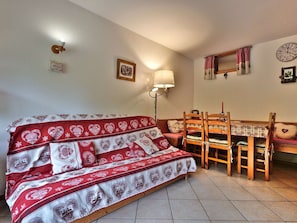 A cozy room with a sofa, a dining table with chairs, a wall clock, and a wall-mounted lamp.
