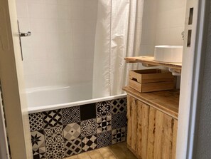 A bathroom with a bathtub, a shower curtain, a patterned tile wall, and a wooden shelf with a box. - Cozy studio in Samoëns center, pets allowed (Samoëns)