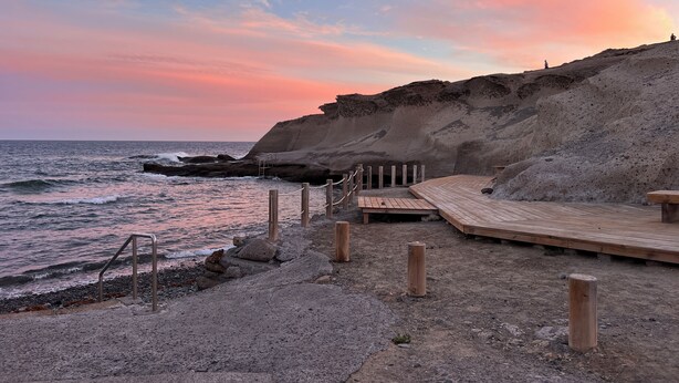 Una terraza de madera con barandilla metálica que da a una playa rocosa y al océano.