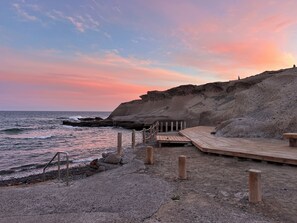 Una terraza de madera con barandilla metálica que da a una playa rocosa y al océano.