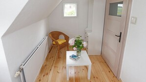 Un pequeño y luminoso dormitorio en ático, con techo inclinado, una silla de mimbre con cojín amarillo, una mesa blanca con un libro y un jarrón de flores, y una puerta de madera.