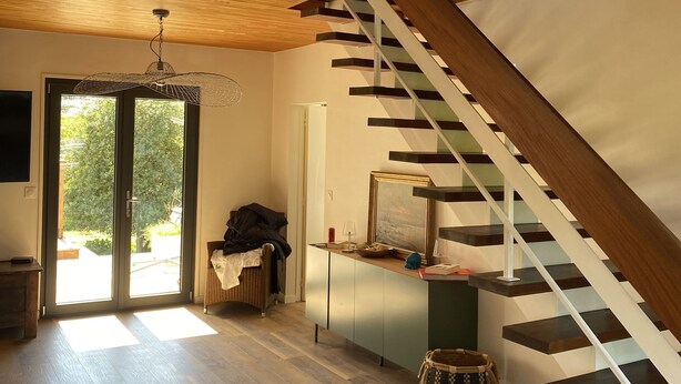 A spacious room with a wooden floor, a staircase with a wooden railing, and a large window allowing natural light.