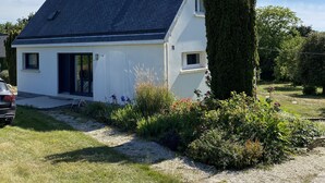 A white house with a dark roof, surrounded by a gravel driveway and a garden with various plants and flowers.