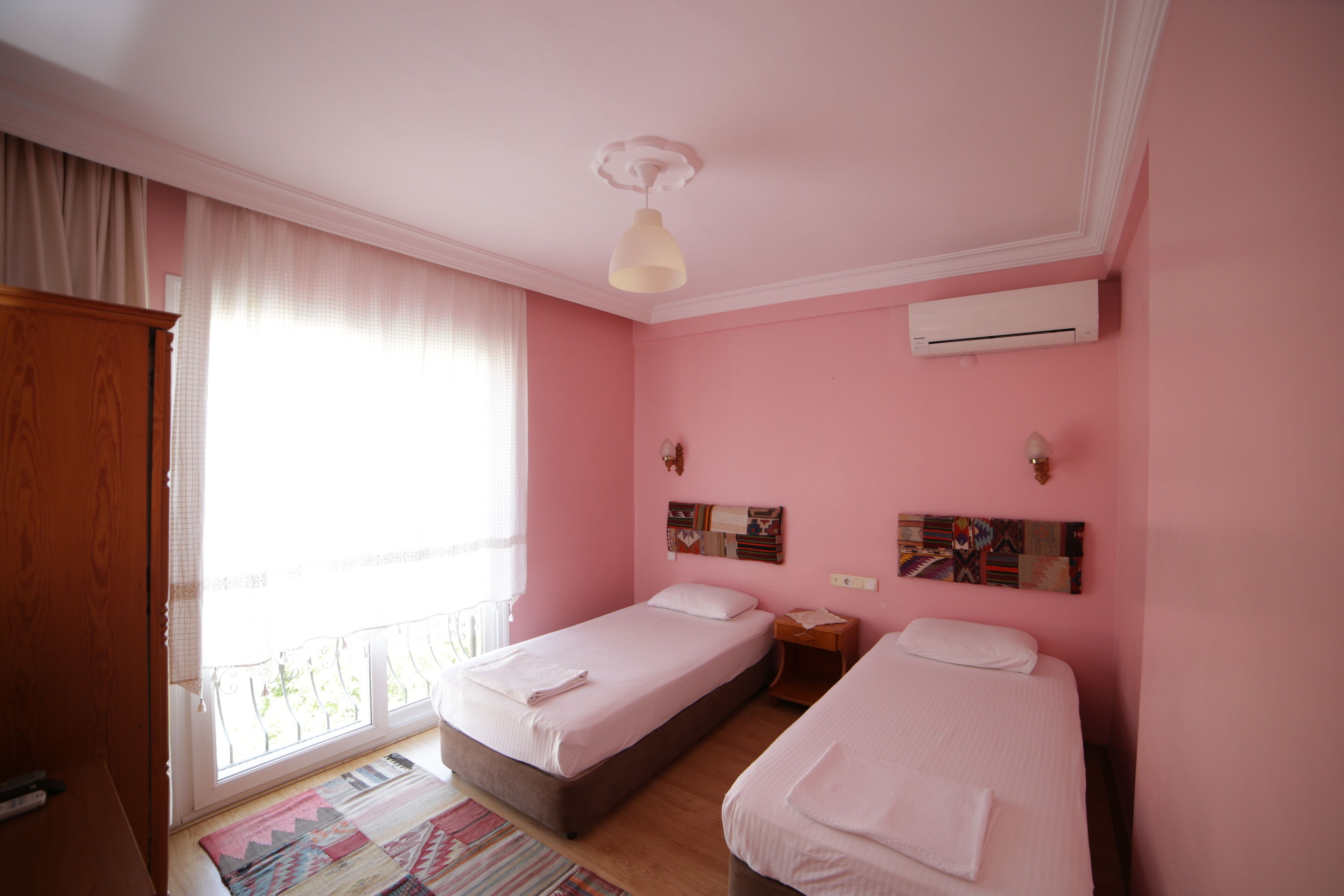 A pink-themed bedroom with two single beds, a wooden wardrobe, a window with curtains, and an air conditioning unit.
