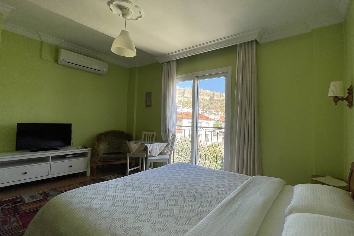 A hotel room with a bed, a television on a dresser, a chair, a table, and a balcony with a view of buildings.