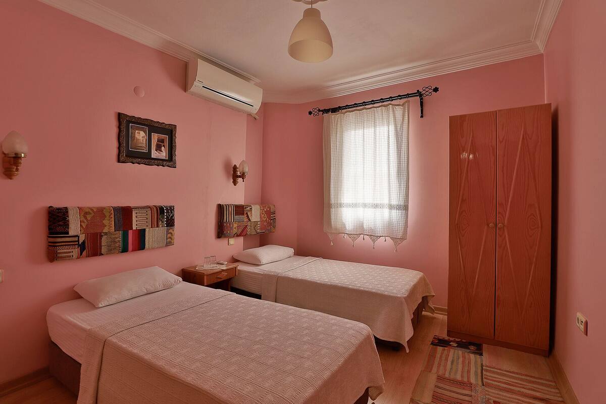 A bedroom with two beds, a wooden wardrobe, a window with lace curtains, and a wall-mounted air conditioner.