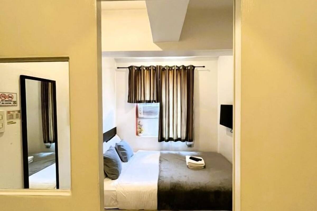 A bedroom with a bed, a bedside table, a mirror, and a window with curtains.