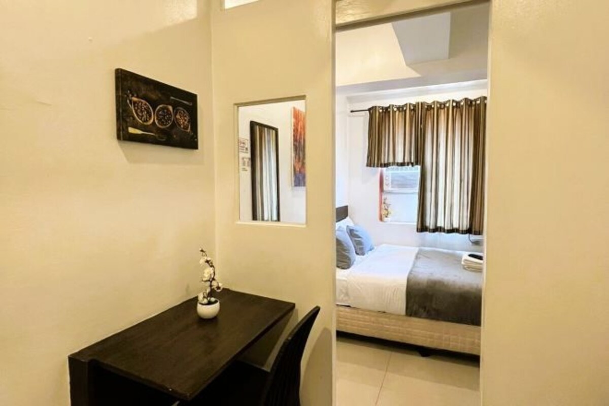 Una habitación pequeña con una cama, un escritorio y una silla. Hay una ventana con cortinas, un cuadro colgado en la pared y una pequeña estatua sobre el escritorio.
