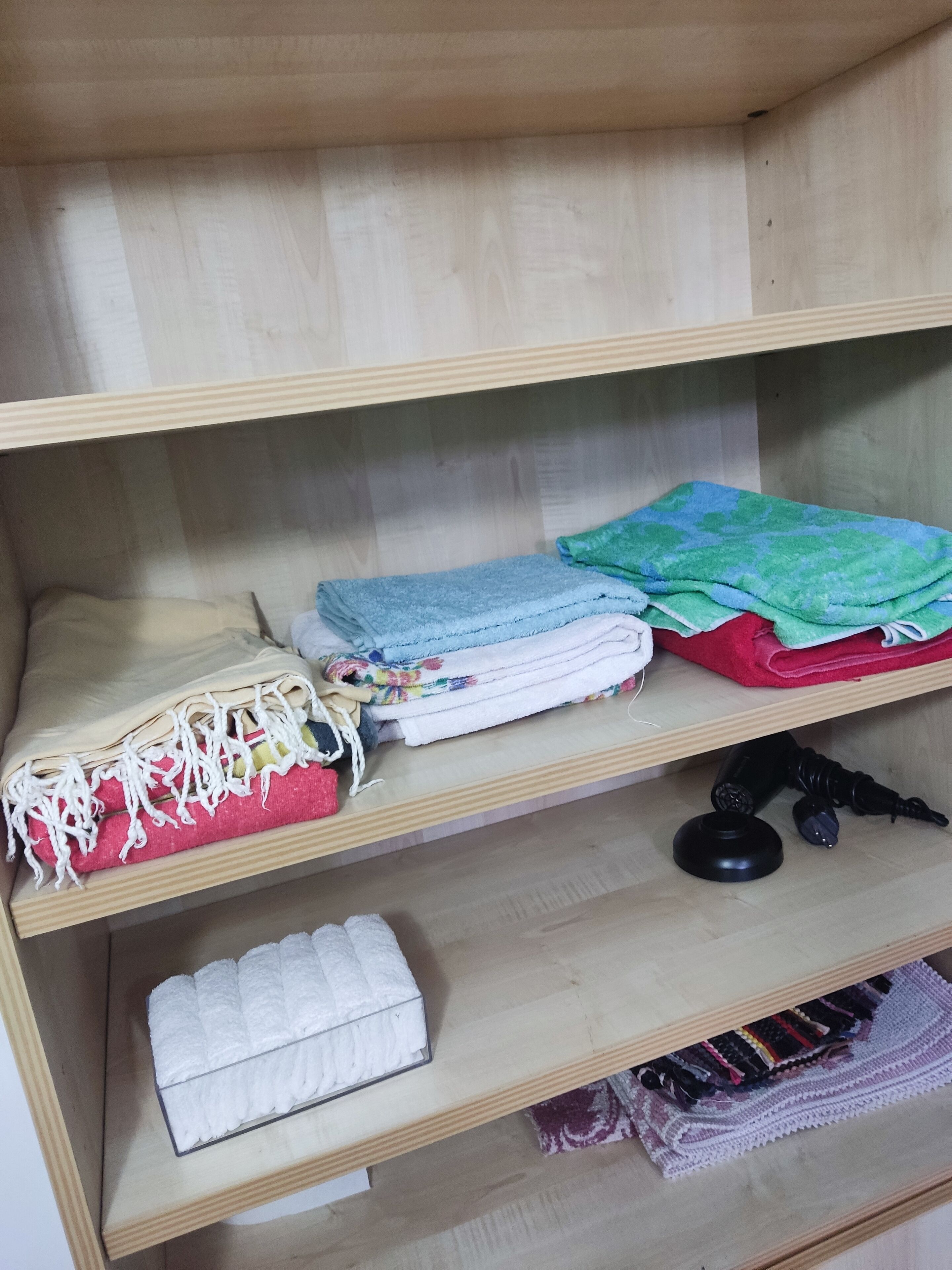 Un estante de madera con toallas dobladas, una caja pequeña de pañuelos y un objeto negro en el estante inferior.