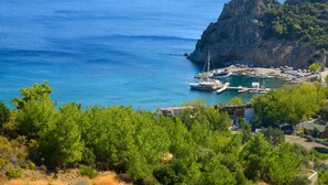 A coastal area with a rocky outcrop, boats docked at a marina, and dense green vegetation in the foreground. - HAYITBÜKÜ AHŞAPEVLERİ (Datça)