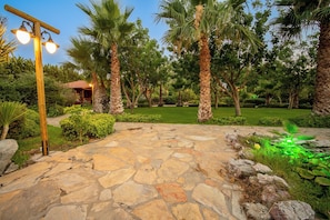 A stone pathway leading to a building with a red roof, surrounded by palm trees and greenery.